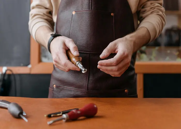 A craftsman wearing a brown leather apron holds a leatherworking tool while working at a wooden workbench with various tools scattered around