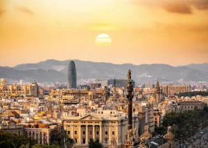 Skyline of Barcelona at sunset, featuring the Columbus Monument, historic buildings, and the modern Torre Glòries with a backdrop of mountains