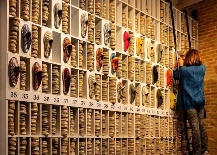 A vendor climbing a ladder to reach espadrilles displayed on a tall, organized wall in a boutique shoe store.