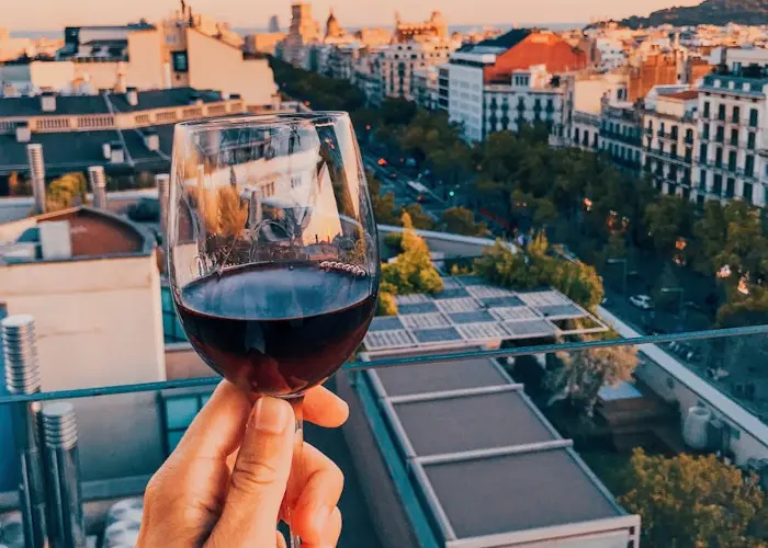 A hand holding a glass of red wine on a rooftop terrace overlooking the cityscape of Barcelona at sunset