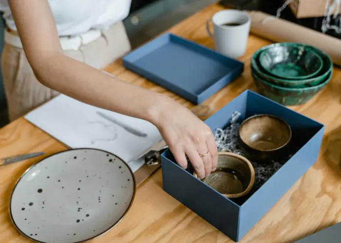 A woman carefully placing handmade ceramic bowls into a blue gift box on a wooden table, surrounded by other pottery and packaging materials.
