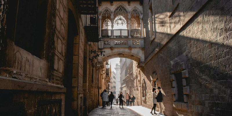 Gothic Quarter in Barcelona with people walking under the iconic neo-Gothic bridge, surrounded by historic stone buildings.