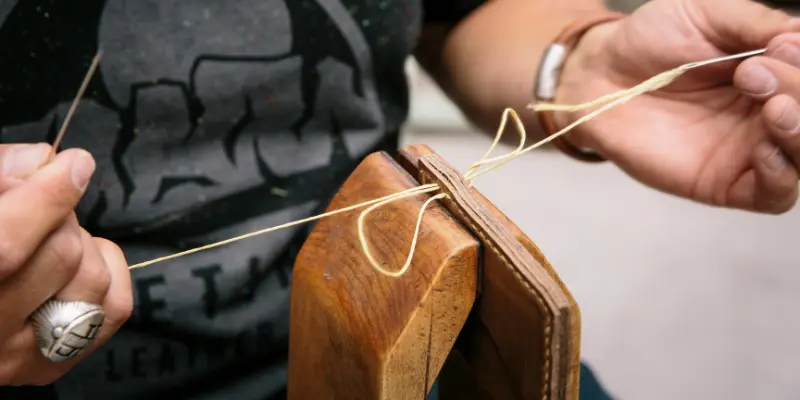 Close-up of an artisan hand-stitching a leather piece in a wooden clamp, pulling waxed thread with two needles