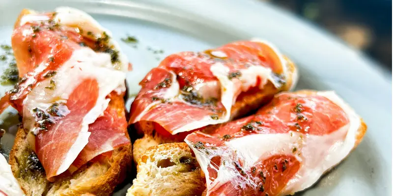 Slices of Jamón Ibérico served on toasted bread with olive oil and herbs on a white plate.