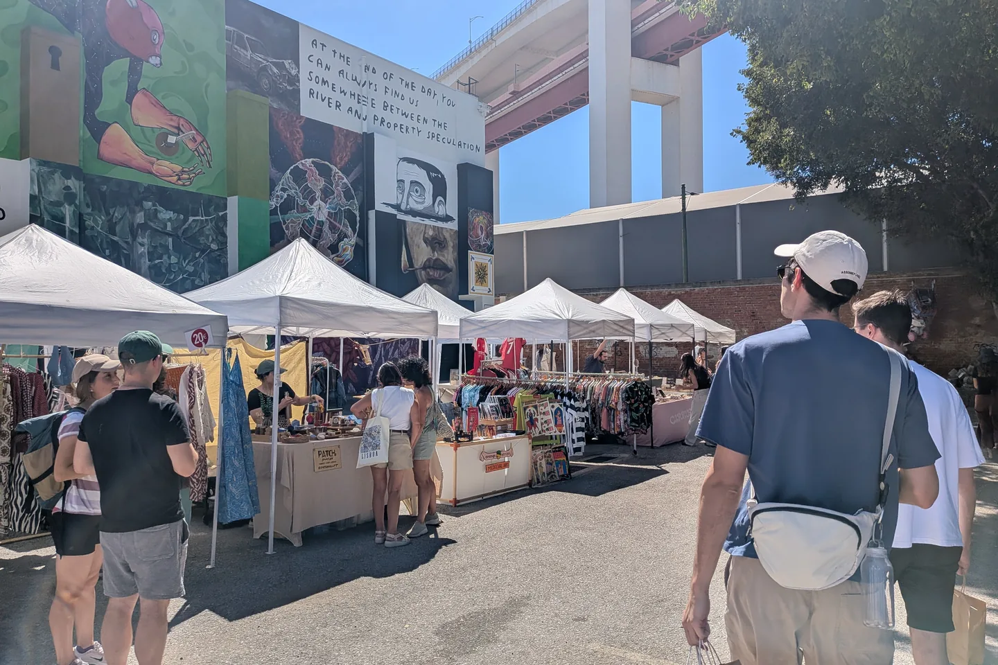 Visitors walking between craft stalls at LX Market with street art and the 25 de Abril Bridge behind.