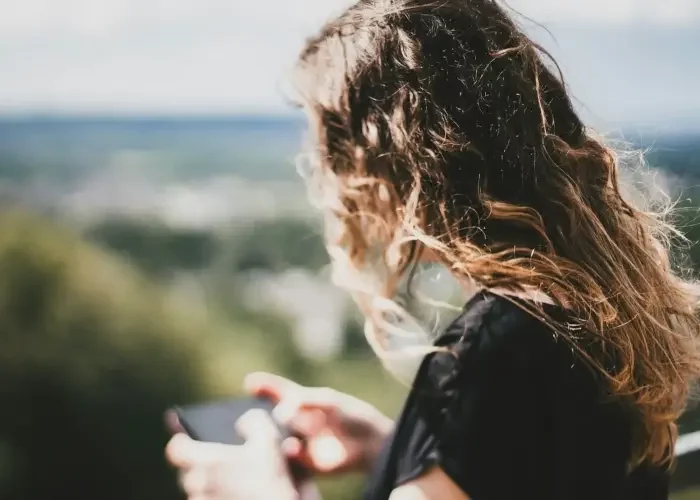 woman-smartphone A woman with wavy brown hair, wearing a black top, holding a smartphone while looking at a blurred outdoor landscape in the background